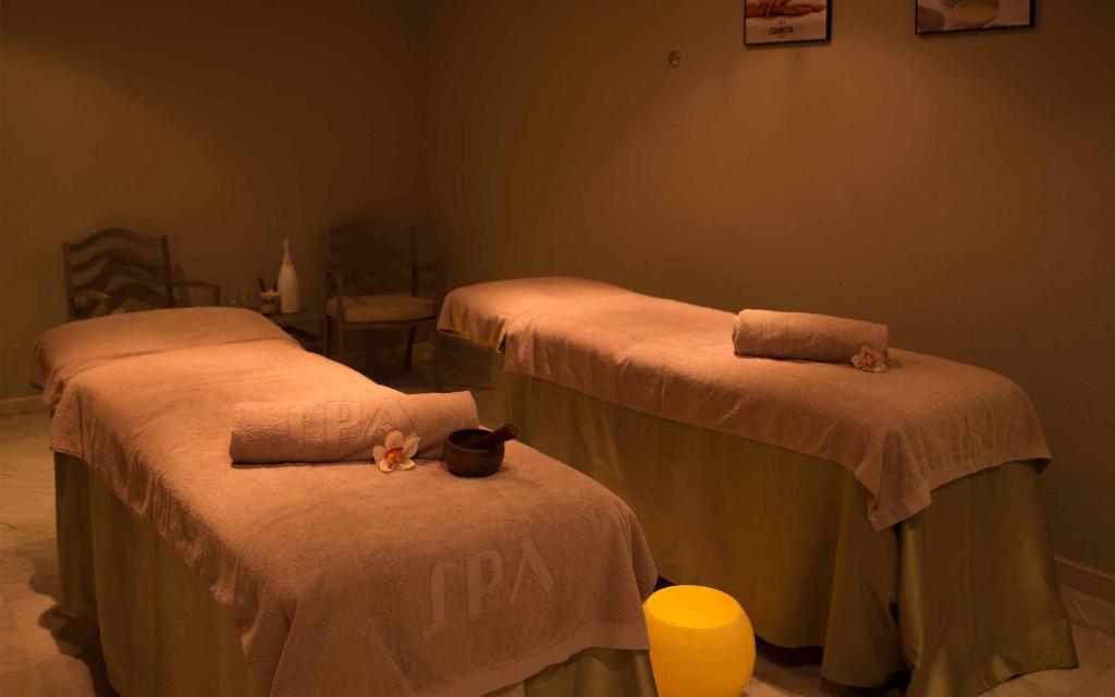 Relaksējoša spa telpa ar masāžas galdiem, pārklātiem ar dvieļiem un ziedu akcentu. Telpas apgaismojums ir maigs un mierīgs, radot patīkamu atmosfēru.