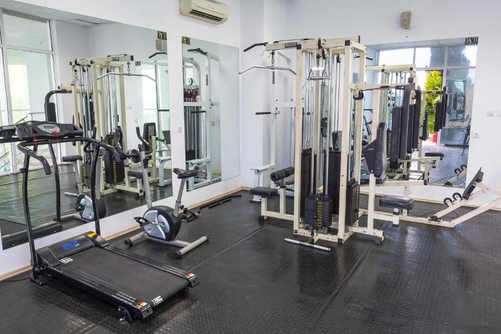 Mūsdienīgs sporta zāles interjers ar dažādām trenažieriem, tostarp skrejceliņu un velotrenažieri, kas atspoguļojas spoguļos. Telpa ir gaiša un plaša, radot motivējošu atmosfēru.