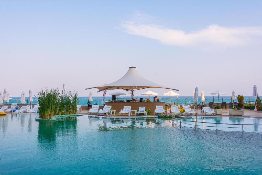 Peldbaseins ar saulessargiem un atpūtas krēsliem, blakus jūrai, ar bāru zem lielas nojumes, radot relaksējošu atmosfēru.