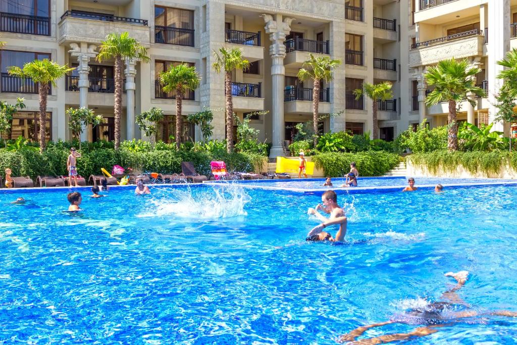 Peldbaseins ar cilvēkiem, kas izbauda sauli un ūdeni, apkārt aug palmām un zaļumiem. Aktīvās atpūtas mirkļi, kur bērni un pieaugušie peld un spēlējas.