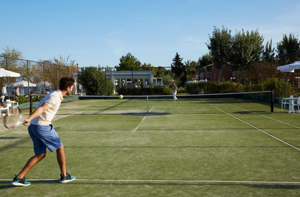 Tenisa spēlētāji spēlē uz laukuma ar zaļu segumu, kamēr saulaina debesis un koki veido patīkamu fonu.