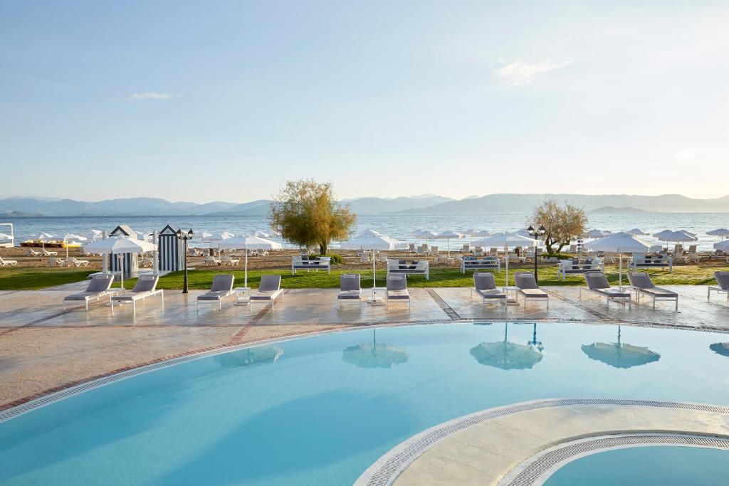 Lielisks skats uz baseinu un pludmali, kur saulaina diena aicina atpūsties uz sauļošanās krēsliem. Fons veido mierīgu jūru un kalnus, radot relaksējošu atmosfēru.