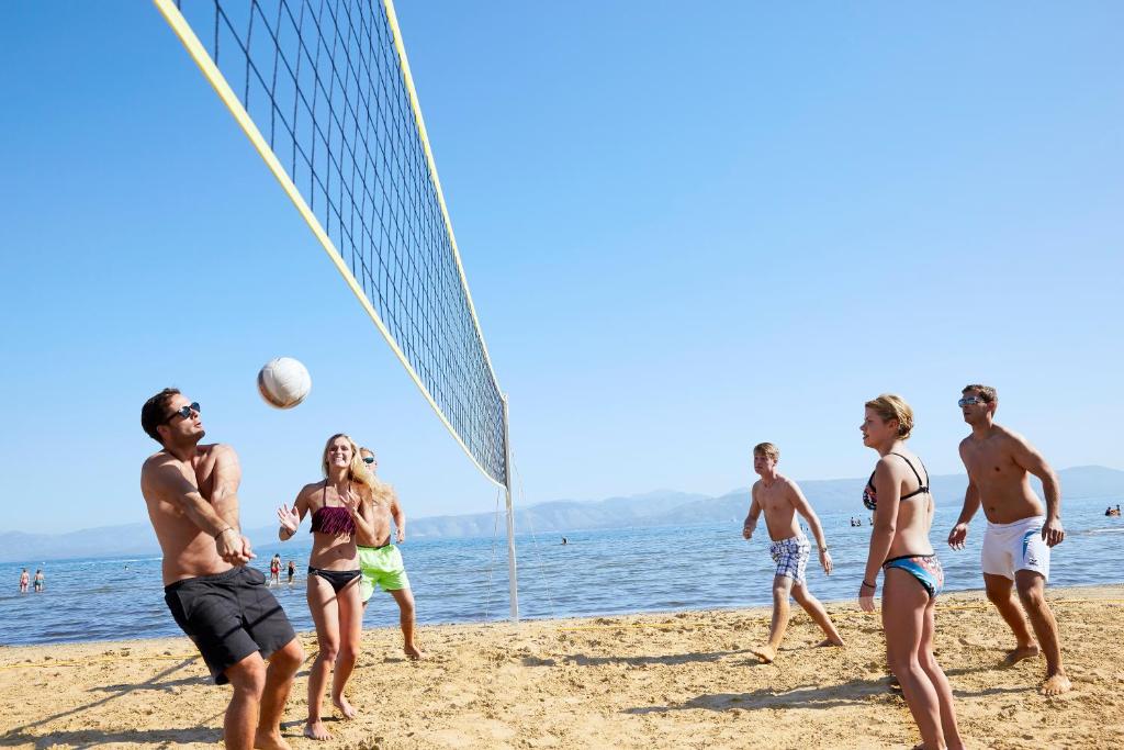 Pludmales volejbola spēle saulainā dienā, kur piedalās vairākas personas. Fons ir jūra un skaidrs debesis.