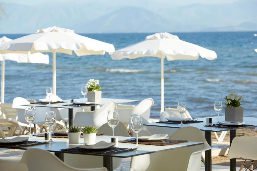 Pludmales restorāna terase ar baltām saulessargiem un galdiem, kas vērsti uz jūru, radot relaksējošu atmosfēru. Jūras skats un harmoniska vide nodrošina ideālu vietu maltītēm.