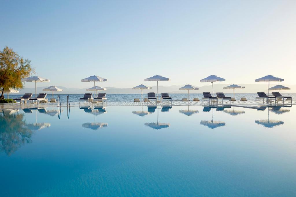 Mūsdienīgs baseins ar sauļošanās krēsliem un saulessargiem pie jūras, atspoguļojot mierīgu ainavu un skaidru debesi. Ideāla vieta atpūtai un relaksācijai.