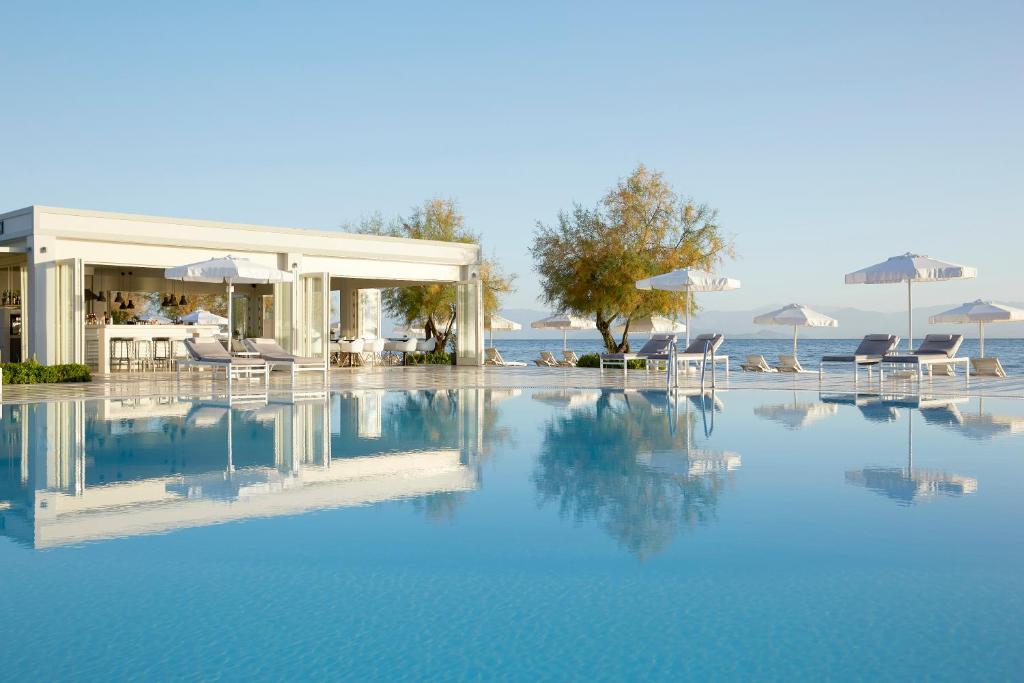 Mūsdienīgs baseins ar baltiem sauļošanās krēsliem un lietussargiem, blakus stilīgai bāra ēkai ar skatu uz jūru. Ideāla vieta atpūtai un relaksācijai.