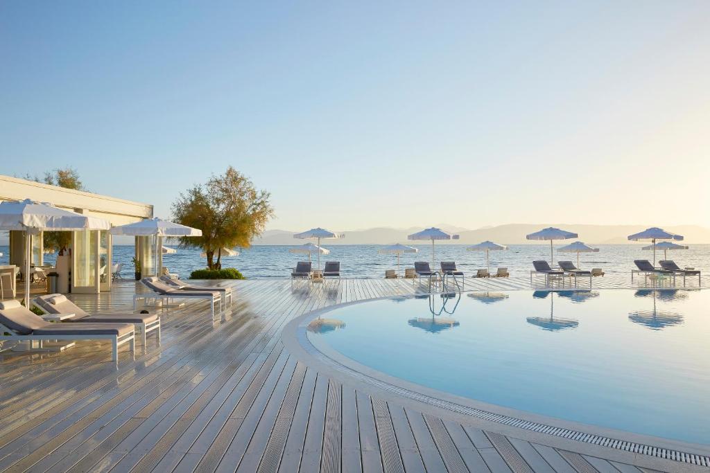 Luksusa atpūtas vieta pie jūras ar peldbaseinu un sauļošanās krēsliem, kas izvietoti uz koka terases, ar skatu uz mierīgu ūdeni un kalnu ainavu.