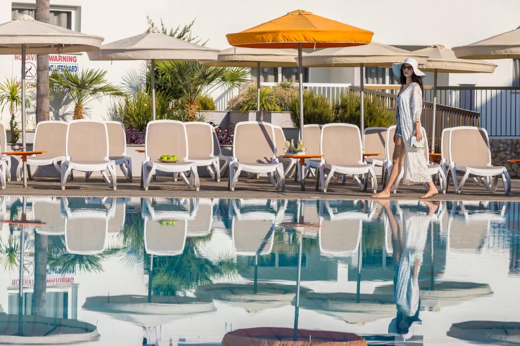 Sieviete, tērpta vieglā apģērbā un platmalē, pastaigā gar baseinu, kurā atspoguļojas sauļošanās krēsli un palmas. Fons ir relaksējoša atpūtas zona ar saulessargiem.