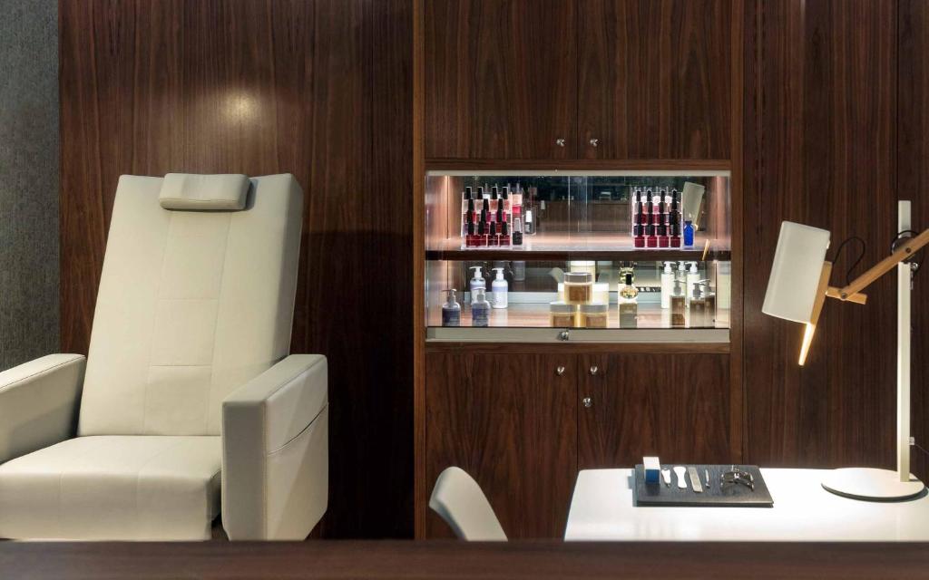 Mūsdienīgs skaistumkopšanas salons ar balto krēslu un elegantu koka apdari, kurā redzami dažādi ādas kopšanas produkti vitrīnā. Galda virsma ir apgaismota ar modernu lampu.