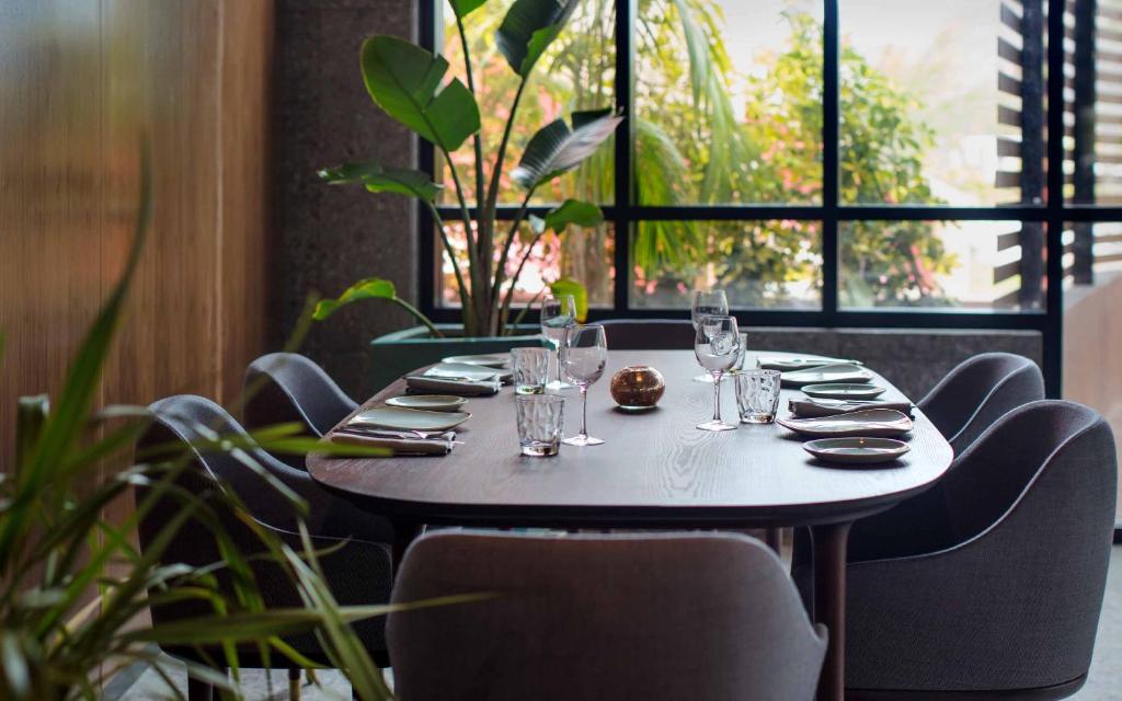Elegants restorāna galds ar moderniem krēsliem, dekorēts ar stikla traukiem un ziediem, norāda uz patīkamu atmosfēru. Lielie logi ļauj ieplūst dabiskajai gaismai, radot siltu un viesmīlīgu vidi.
