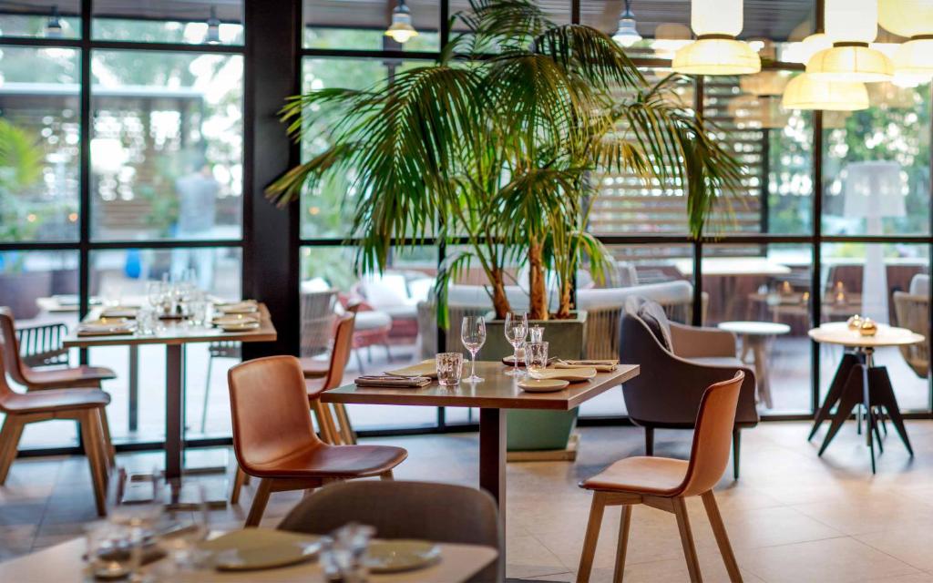 Mūsdienīga restorāna iekārtojums ar zaļām palmām, elegantiem galdiem un komfortablām krēsliem. Lielie logi nodrošina dabisku apgaismojumu un skatu uz ārpusi.