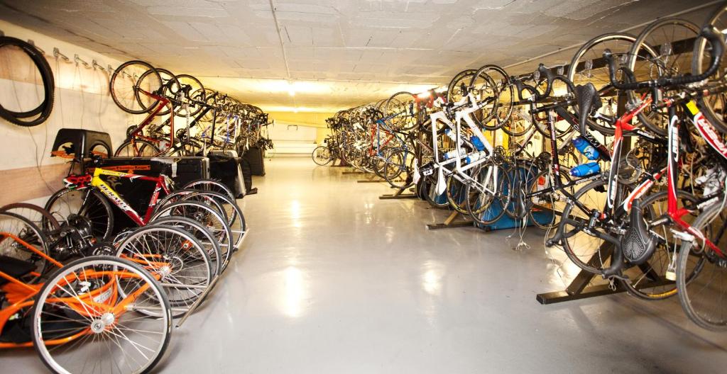 Velosipēdu noliktava ar vairākiem stāvošiem un guļošiem velosipēdiem, kas nepieciešami ikdienas lietošanai. Telpa ir labi organizēta un apgaismota.