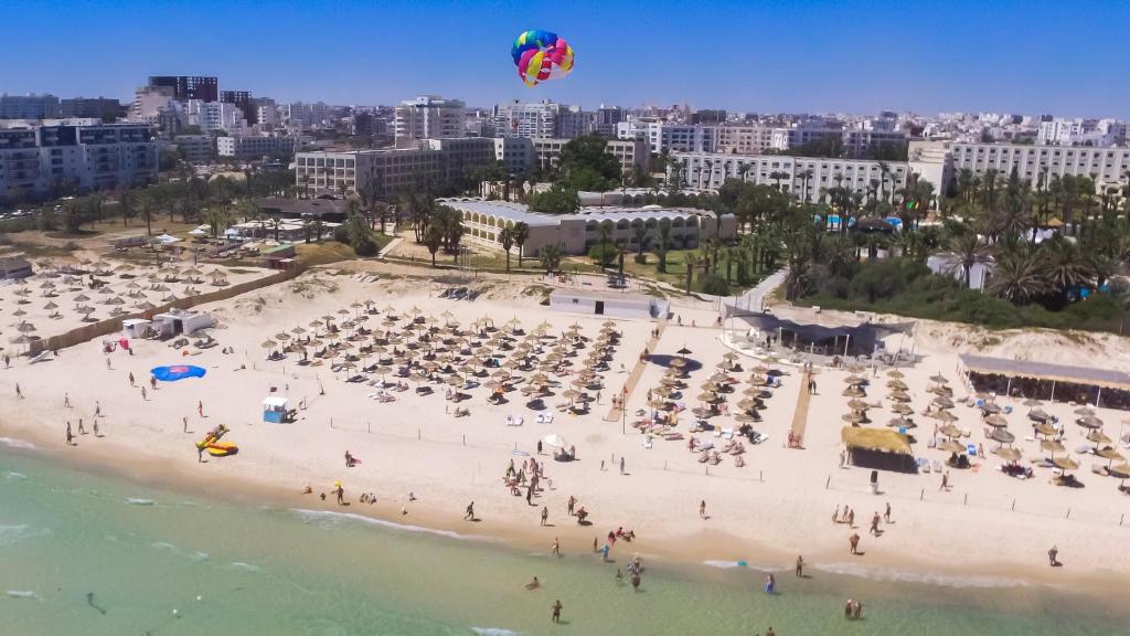Pludmales skats ar cilvēkiem, kas atpūšas, saulessargiem un paragliding lidotāju. Fons ietver pilsētas ainavu ar modernām ēkām un zaļām palmām.