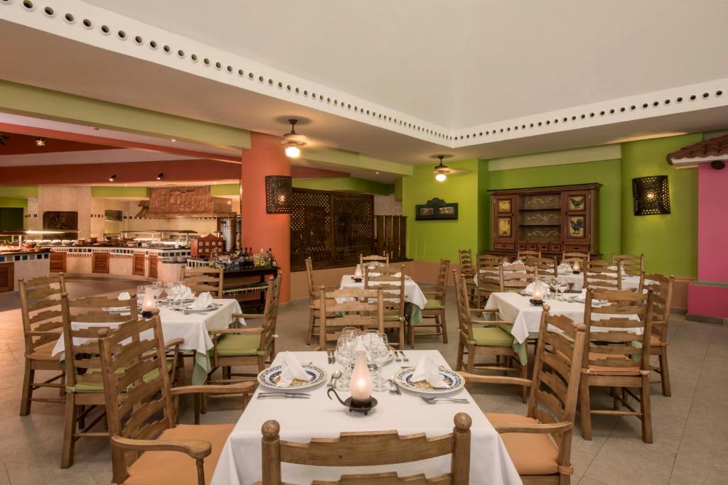 Krāsains restorāna interjers ar koka galdiem un krēsliem, klātiem ar baltām galdautām. Redzama atvērta virtuve un dekoratīvi elementi uz sienām.