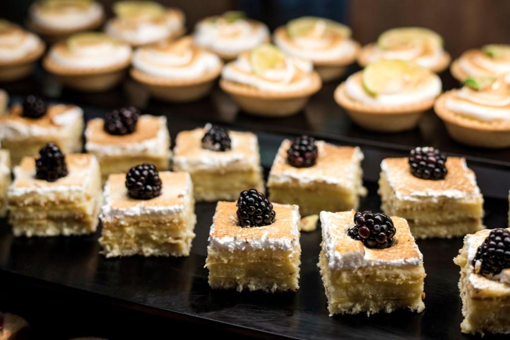 Saldēti kūku gabali ar ogām un krēmīgu virsu, blakus maziem kūciņām ar citrona krēmu un dekoratīvām garnējumiem.