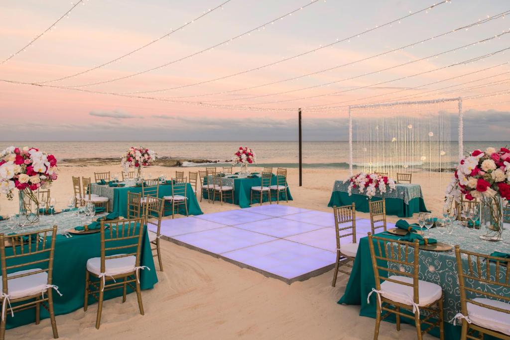 Eleganta pludmales kāzu vieta ar zilo galdautu, grezniem ziedu kompozīcijām un apgaismojumu virs galdiem. Skaista jūras ainava un romantiska atmosfēra.