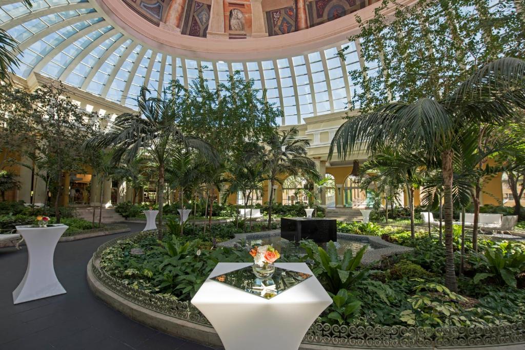 Labi apzaļumota iekštelpa ar stikloto jumtu, kurā ir balti galdi ar ziediem, apstādījumi un palmām. Gaisīgs un elegants dizains, ideāls svinībām.