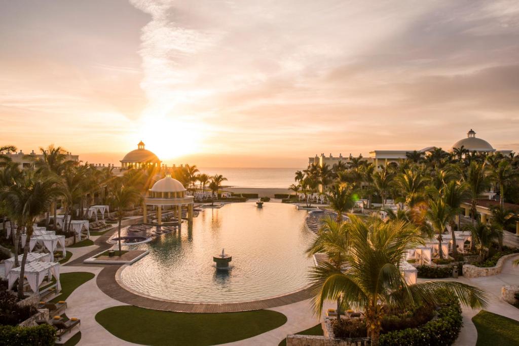 Viesnīcas kompleksa ainava pie pludmales ar palmām un saulrietu fona. Izteikta mierīga atmosfēra ar peldbaseinu centrā un elegantiem atpūtas laukumiem.