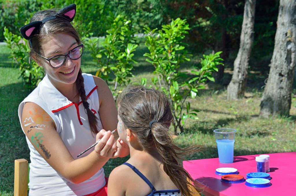 Bērnu sejas krāsošana vasaras pasākumā, kur krāsotājs palīdz meitenei ar krāsu. Apkārt zaļa vide un krāsaini krāsas trauki uz galda.