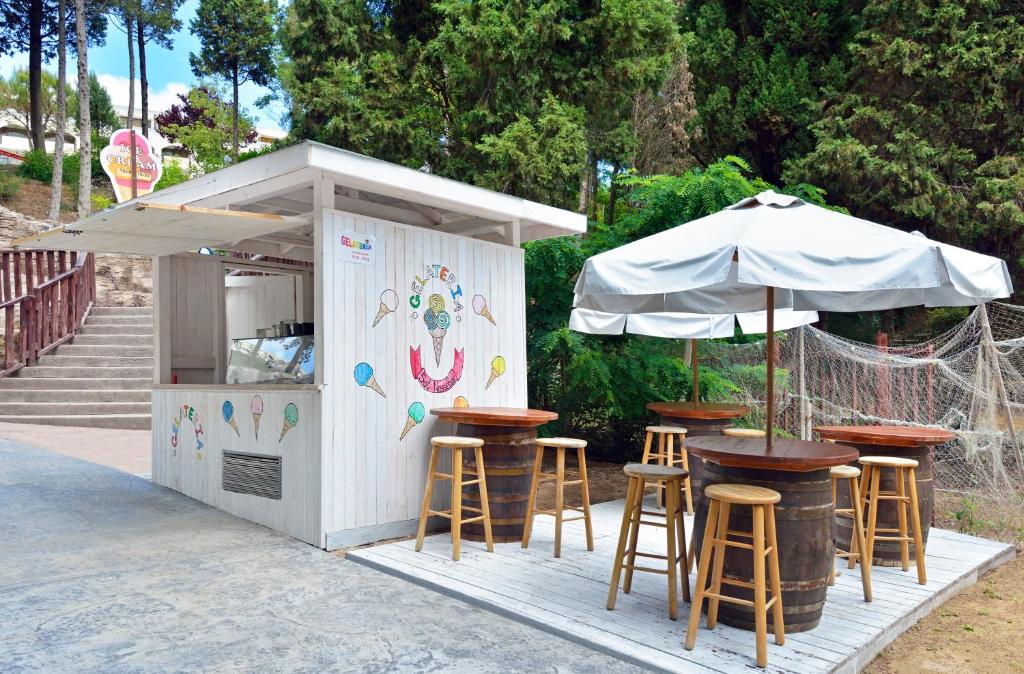 Apsildāms saldējuma kiosks ar galdiem un krēsliem, iedvesmots no jūras tematikas, atrodas zaļā vidē. Kiosks piedāvā dažādus saldējuma veidus un ir ideāla vieta vasaras atpūtai.