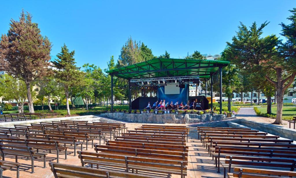 Atklāta amfiteātra skatuve ar koka soliem, apzaļumota vide un saulaina debesis. Ideāli piemērota kultūras pasākumiem un koncertiem.