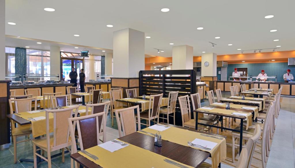 Mūsdienīgs restorāna interjers ar koka galdiem un krēsliem, kas izkārtoti ar dzelteniem galdautiem. Redzami pavāri virtuvē, gatavojot ēdienus.