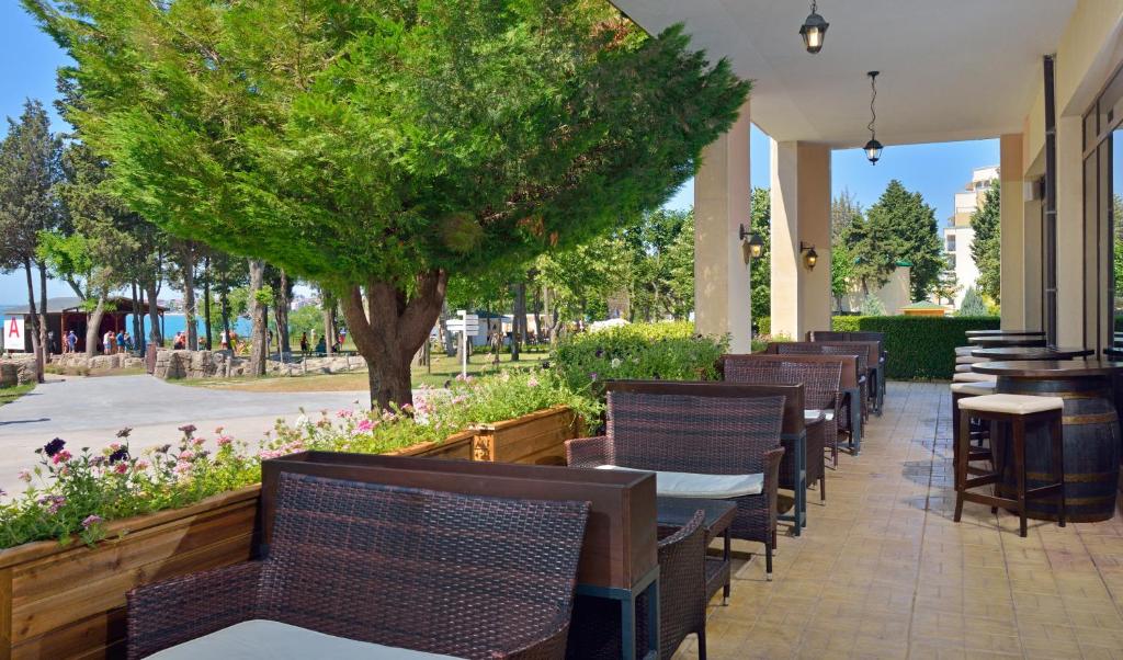 Terases zona ar viesmīlīgām mēbelēm un zaļiem kokiem, kas nodrošina ērtu atpūtu. Fons ietver dārzu, kuru rotā ziedi un tuvumā esošais jūras skats.