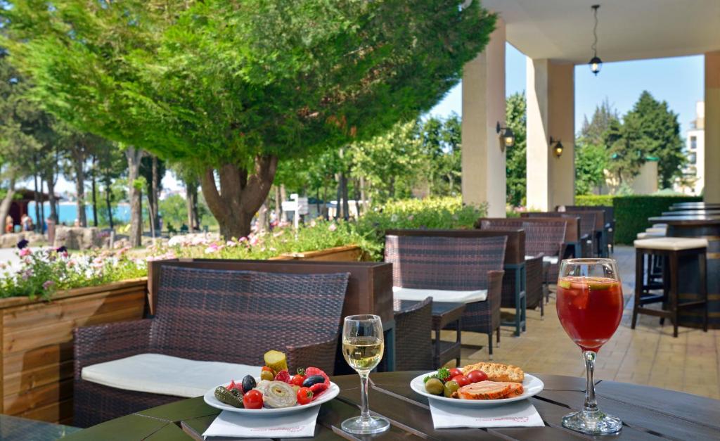 Āra kafejnīca ar zaļiem kokiem un ziediem, kurā redzami divi dzērieni un ēdieni uz galda, aicina uz relaksējošu maltīti. Ideāla vieta, lai izbaudītu garšīgu ēdienu un dzērienus brīvā dabā.