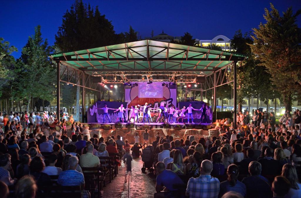 Nakts izrāde uz skatuves, apgaismota ar krāsainām gaismām, pulcē skatītājus, kas bauda deju un izpildījumu ārā.