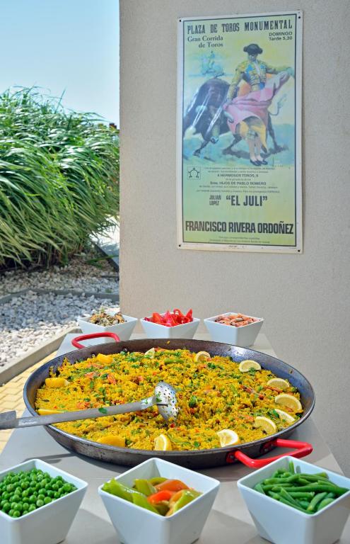 Tradicionāla spāņu paelja ar dārzeņiem un citronu šķēlītēm, novietota uz galda blakus dekoratīvai plakātam par koridu. Apkārt paeljai ir dažādi dārzeņi un salāti.