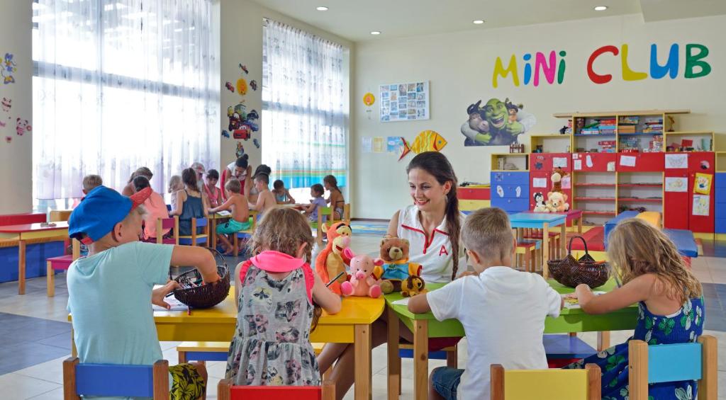 Bērni, kas piedalās aktivitātēs Mini klubā, priecīgi strādā pie galdiem, kamēr pieaugušais uzrauga un iesaistās. Telpa ir gaiša un krāsaina, ar rotaļlietām un zīmējumiem uz sienām.
