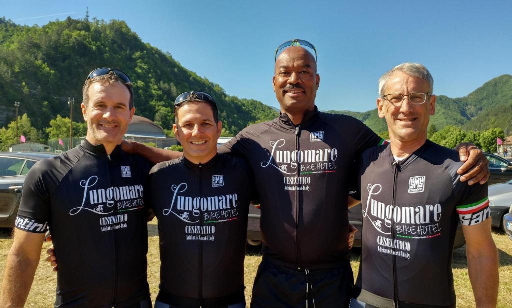 Četri vīri, tērpušies sportiskās komandas tērpos ar "Lungomare Bike Hotel" logo, pozē kopā kalnu vidē, baudot saulainu dienu. Fons ir dabas skats ar kalniem un zilu debesi.