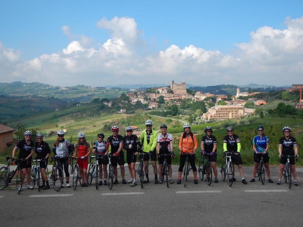 Grupas riteņbraucēju fotogrāfija gleznainā ainavā ar kalniem un mājām fona. Riteņbraucēji pozē, tērpušies sporta apģērbā un ķiverēs, gatavojoties braucienam.