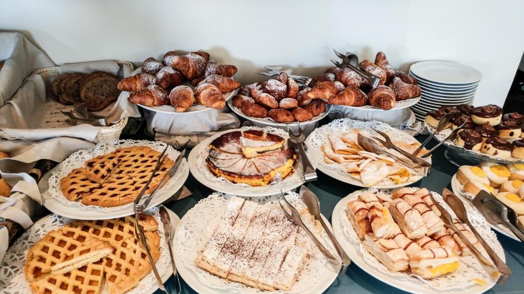 Bagātīgs konditorejas izstrādājumu klāsts, ieskaitot kruasānus, kūkas un vafeles, izlikts uz galda ar dekoratīvām šķīvju malām un metāla karotēm.