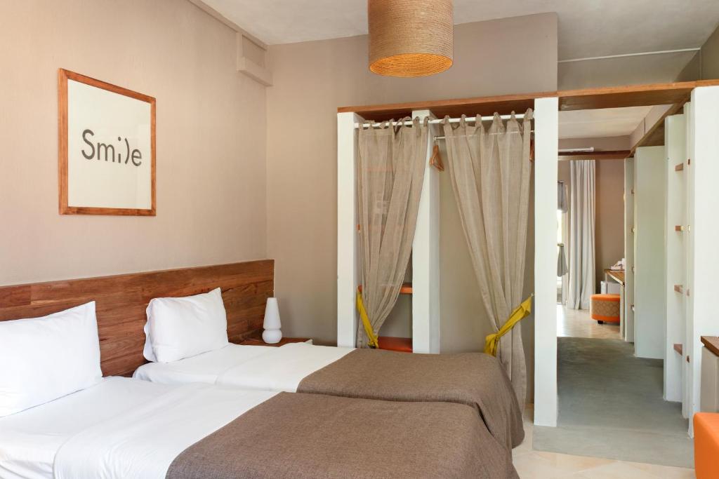 Gaiša viesnīcas numura interjers ar divām gultām, koka elementiem un "Smile" mākslas darbu uz sienas. Numurā ir aizkari un moderni mēbeles, radot mājīgu atmosfēru.