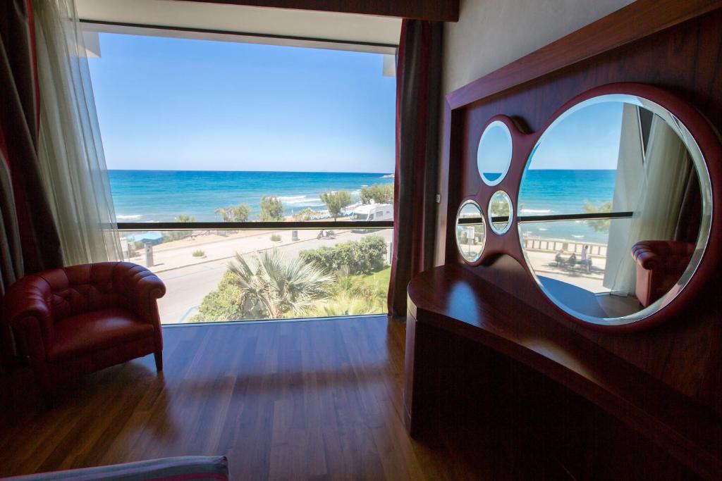 Numura skats uz jūru ar plašu logu un sarkanu ādas krēslu. Moderns interjers ar spoguļiem un koka grīdu, radot viesmīlīgu atmosfēru.