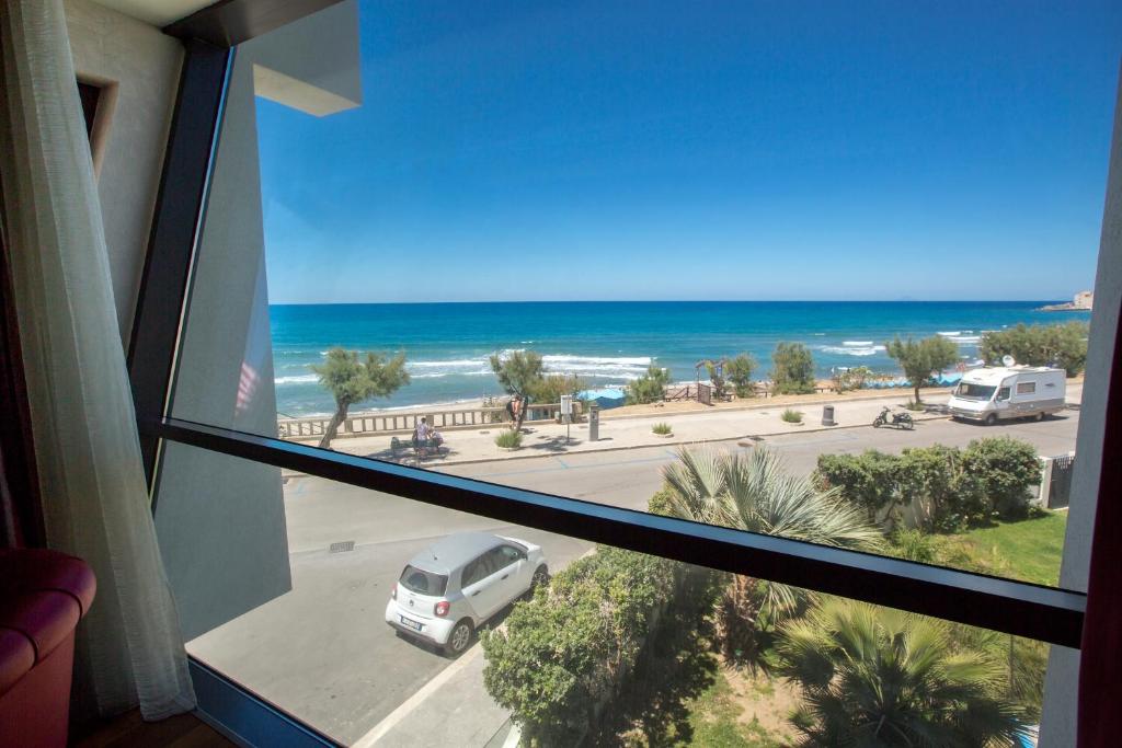 Skats no loga uz pludmali un jūru, ar zilu debesu fonu. Redzami cilvēki, kas pastaigājas gar krastu, un automašīnas uz ceļa.