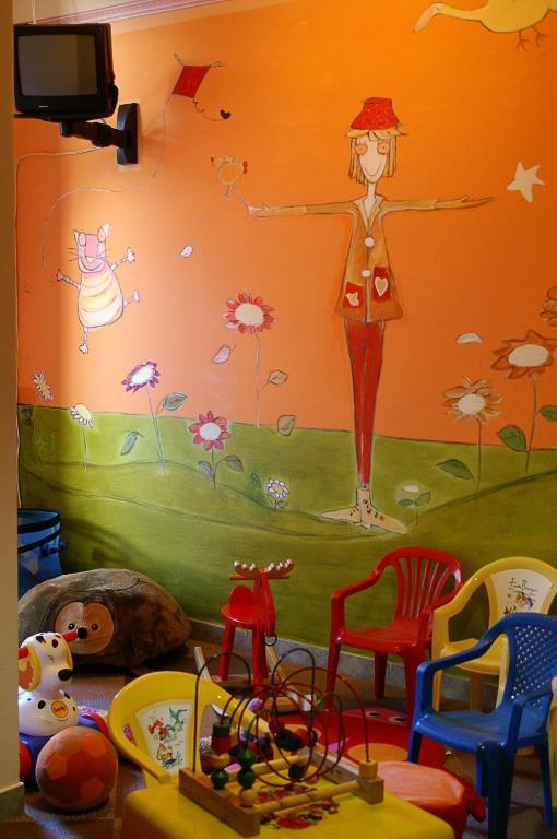 Krāsaina bērnu spēļu istaba ar zīmējumiem uz sienas, kas attēlo priecīgas figūras un ziedus. Telpā redzami dažādi bērnu krēsli un rotaļlietas.