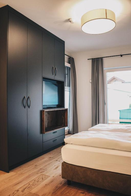 Mūsdienīga guļamistaba ar melnu skapju sienu, iebūvētu televizoru un koka elementiem, kas rada siltu atmosfēru. Logu aizsedz pelēki aizkari, bet uz grīdas ir koka dēļi.