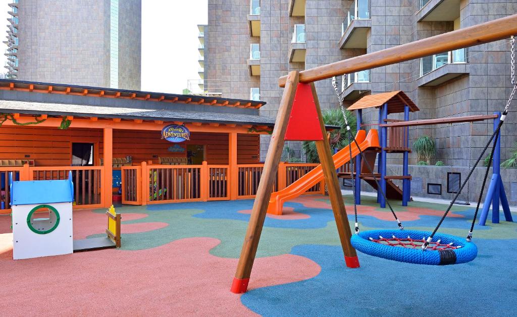 Bērnu rotaļu laukums ar šūpolēm, slidkalniņu un krāsainu grīdu, kas izveidots drošai spēlēšanai. Apkārt redzami moderni daudzdzīvokļu nami.