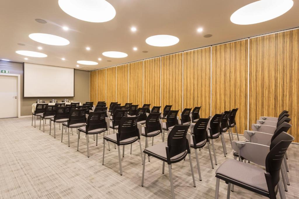 Konferenču telpa ar rindu melnām krēslām un lielu ekrānu, gatava sanāksmēm vai prezentācijām. Sienas apdarinātas ar koka faktūru, radot modernu un profesionālu atmosfēru.