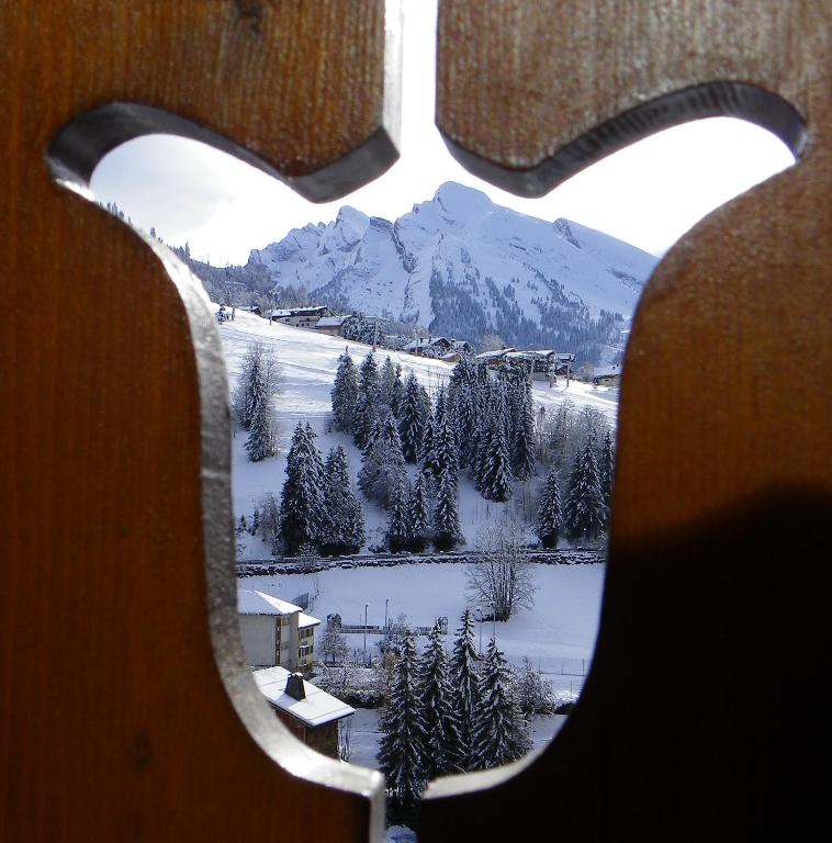 Apsnigušas kalnu ainavas skats caur dekoratīvu koka logu. Sniegotās meža zonas un māju jumti veido mierīgu ziemas ainavu.