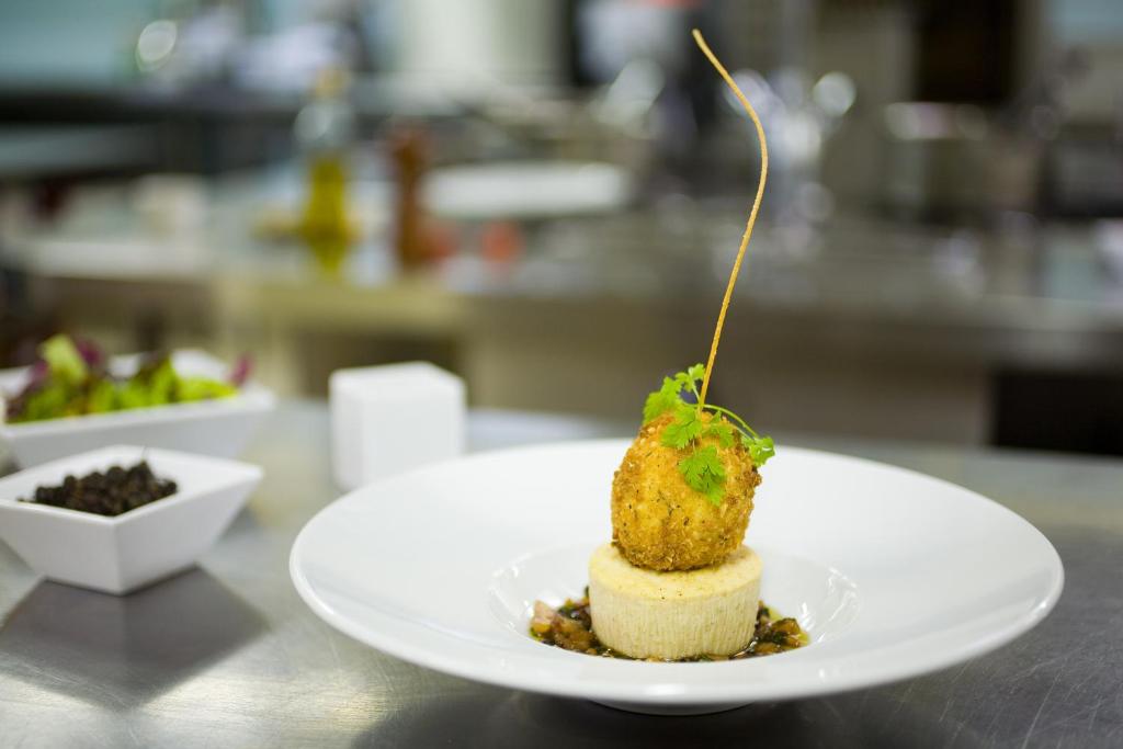 Mūsdienīgs gastronomisks ēdiens ar kraukšķīgu bumbiņu, kas pasniegta uz krēmveida pamatnes un dekorēta ar koriandru. Fonā redzama virtuve ar sastāvdaļām un traukiem.