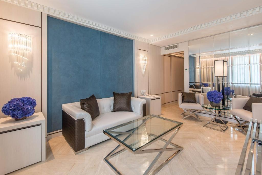 Mūsdienīgs viesistabas interjers ar gaišiem dīvāniem, stikla galdu un zilu akcentu, kas veidots no ziediem. Telpa ir eleganti iekārtota ar gludām līnijām un spoguļotām virsmām.