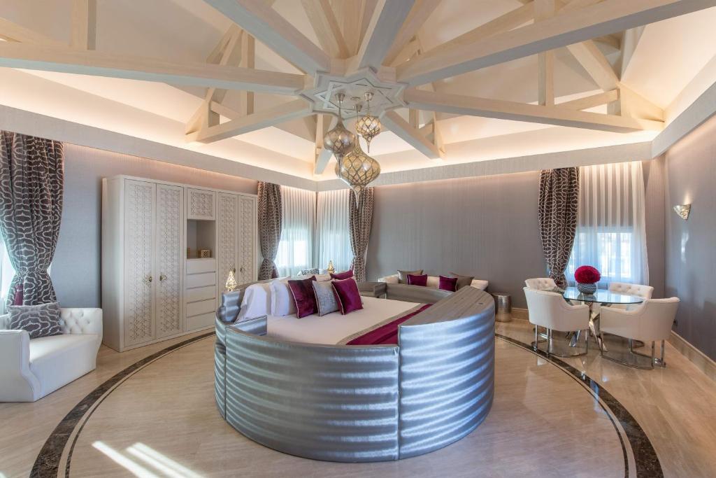 Mūsdienīgs guļamistabas interjers ar apaļu gultu, elegantiem dīvāniem un modernu apgaismojumu, radot greznu atmosfēru. Melanžas toņu aizkari un dekoratīvie elementi piešķir telpai stilu.