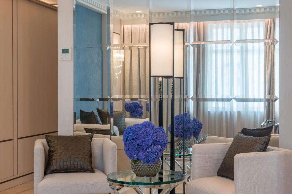 Mūsdienīgs interjers ar baltām krēsliem, stikla galdu un zilu ziedu kompozīciju, papildinātu ar spoguļa sienu un maigu apgaismojumu.