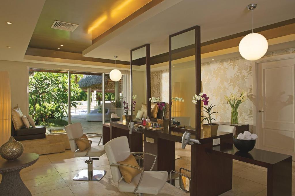 Mūsdienīgs skaistumkopšanas salons ar elegantiem krēsliem, lieliem spoguļiem un zaļo dabu aiz loga, radot relaksējošu atmosfēru. Salons ir aprīkots ar dažādiem skaistumkopšanas produktiem un dekoratīviem augiem.