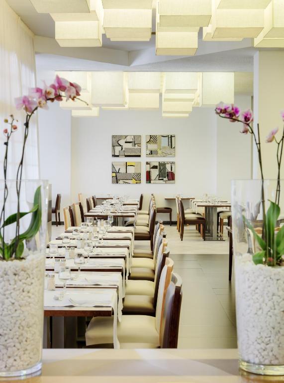 Mūsdienīgs restorāna interjers ar elegantiem galdiem un krēsliem, dekorēts ar orhidejām un mākslas darbiem uz sienām. Gaismas elementi rada siltu atmosfēru, radot patīkamu vidi ēdienreizei.