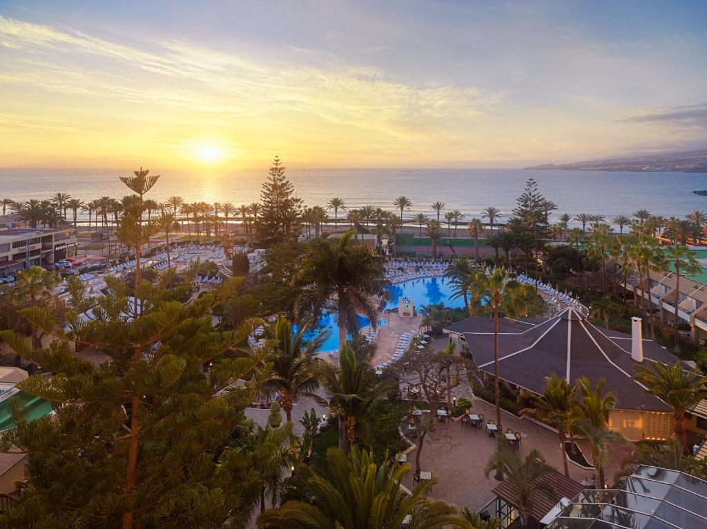 Saulriets virs jūras, redzams viesnīcas baseins un palmas, radot relaksējošu atmosfēru. Ideāla vieta atpūtai pie ūdens.
