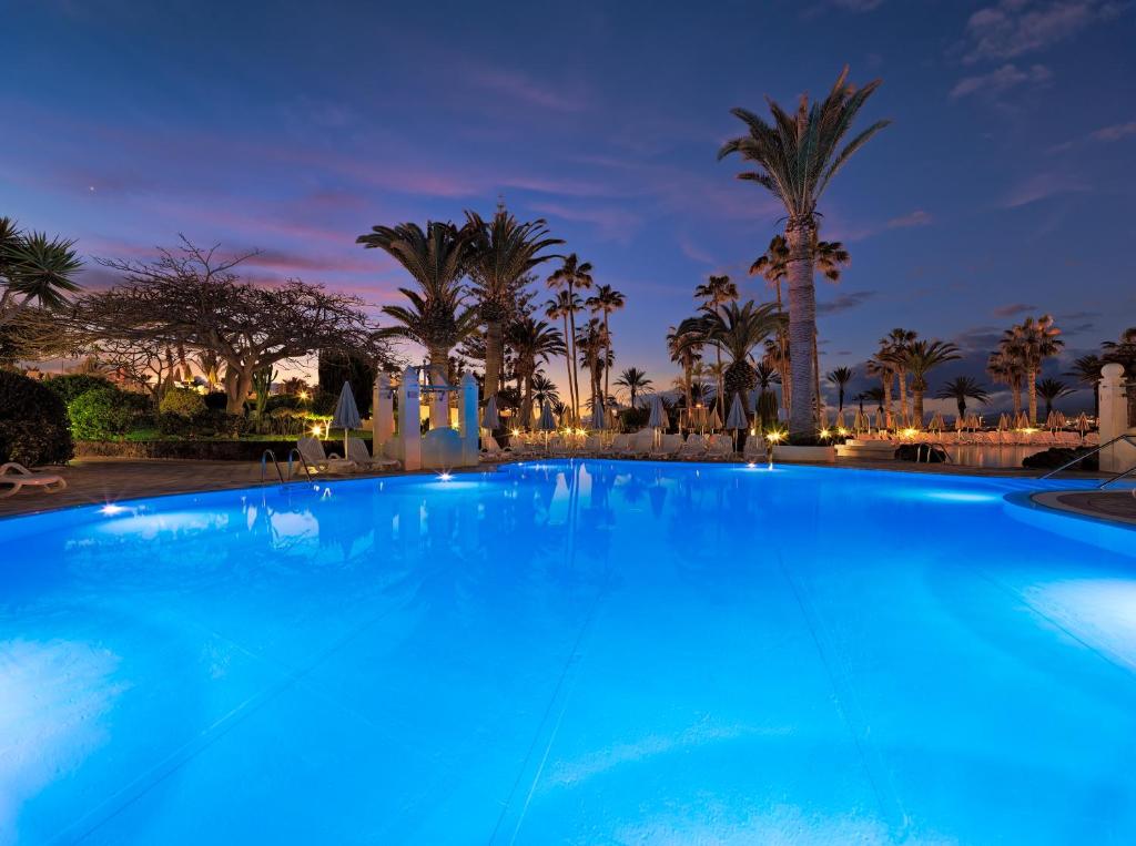 Lieliska nakts ainava ar spožu zilu baseinu, apkārt palmām un izgaismotiem dārziem, radot relaksējošu atmosfēru.